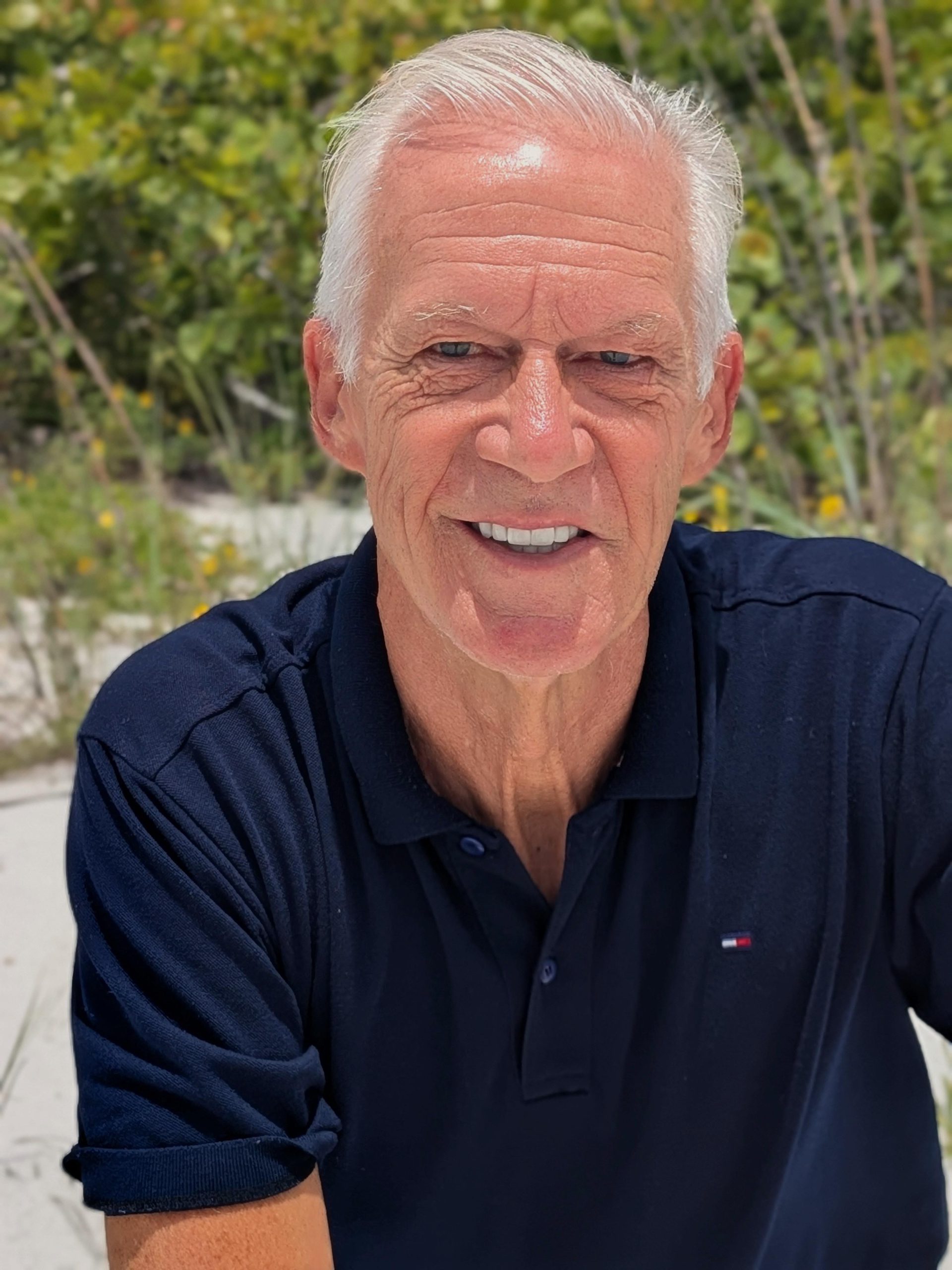 A smiling silver-haired man in a navy polo shirt outdoors on a sunlit beach path.